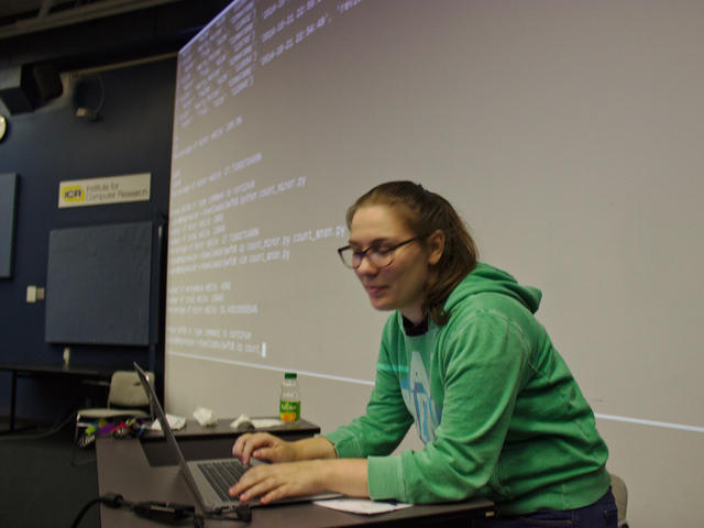 Elana Hashman, typing on a laptop at a podium in front of a screen displaying a command line prompt and Python code